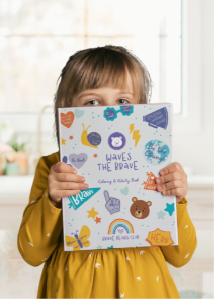 Child holding a colorful book in front of their face with a white wall and window in the background
